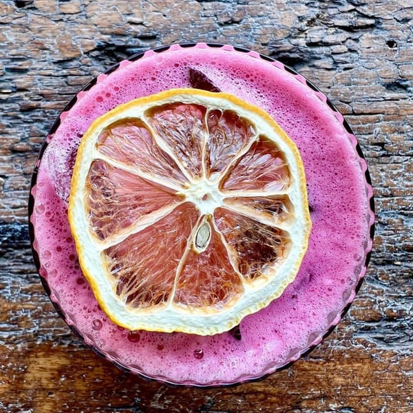 Overhead view of a pink foamy drink garnished with a dried citrus slice on a wooden surface.
