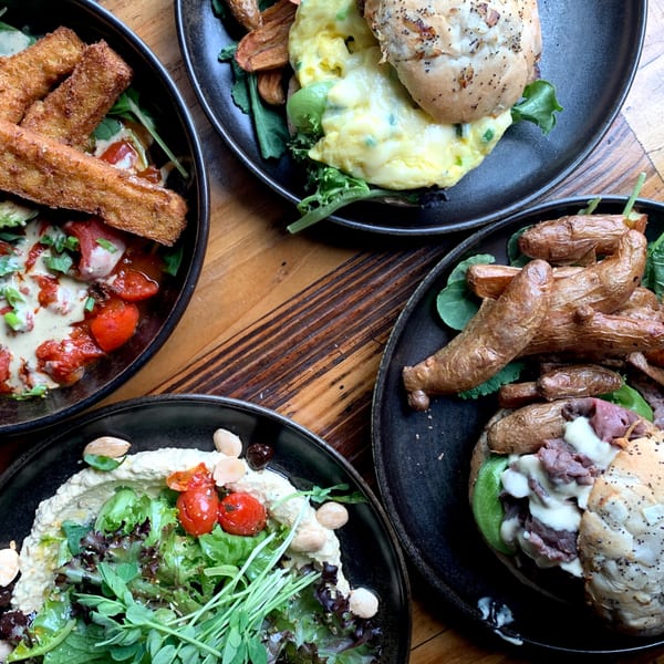 Four plates of Bandaloop's healthy dishes, including sandwiches, hummus, and fried plantains, on a wooden table.
