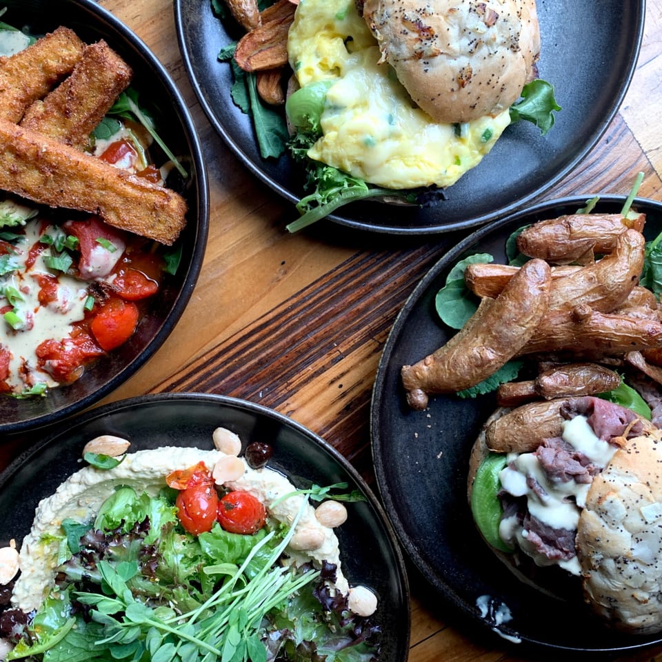 Four plates of Bandaloop's healthy dishes, including sandwiches, hummus, and fried plantains, on a wooden table.