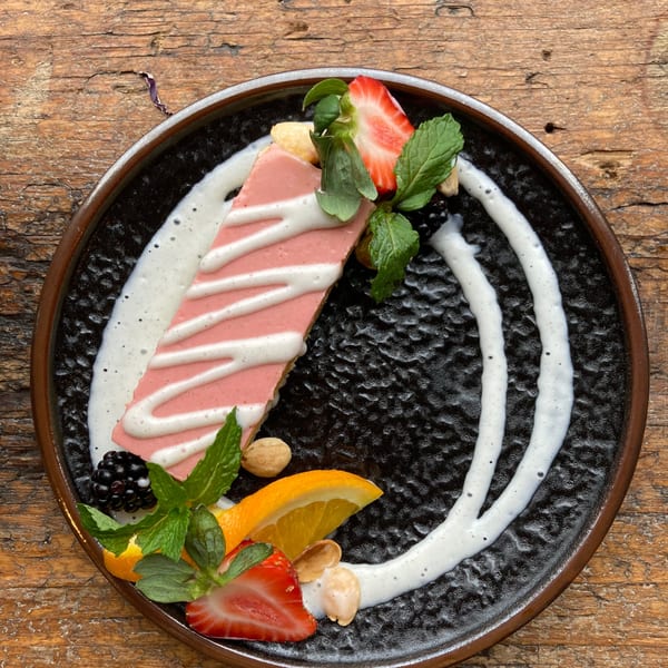 Pink dessert slice with white drizzle, strawberries, blackberries, orange, mint, and nuts on a dark plate.