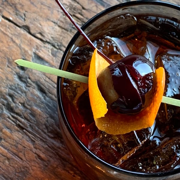 Dark cocktail with ice, garnished with an orange peel and a cherry on a wooden table.
