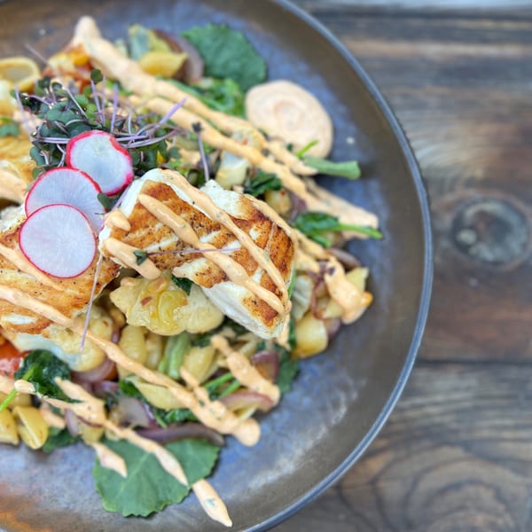 Grilled halloumi with roasted potatoes, greens, radishes, and a creamy sauce in a dark bowl.