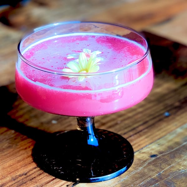 A vibrant pink cocktail in a coupe glass, garnished with a small yellow flower, on a wooden table.